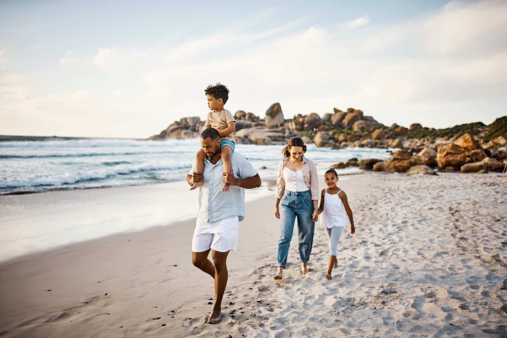 A family of four walking along a sandy beach with scenic rocks in the background, enjoying a relaxed seaside day.